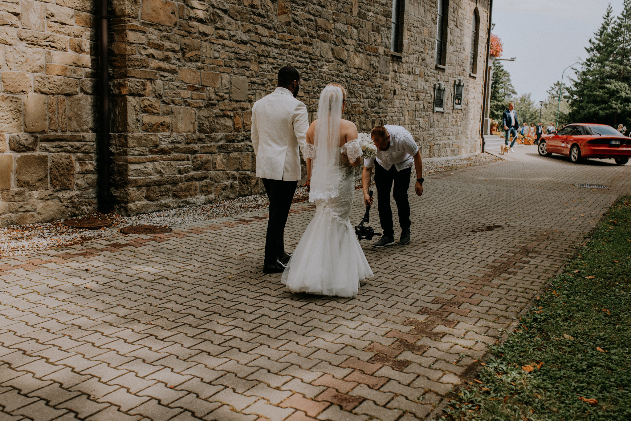 Para młoda w trakcie sesji ślubnej na zewnątrz, mężczyzna w białym smokingu, panna młoda w sukni z odkrytymi ramionami, kamerzysta pochylony nad sprzętem na brukowanej ścieżce, w tle kamienny budynek.