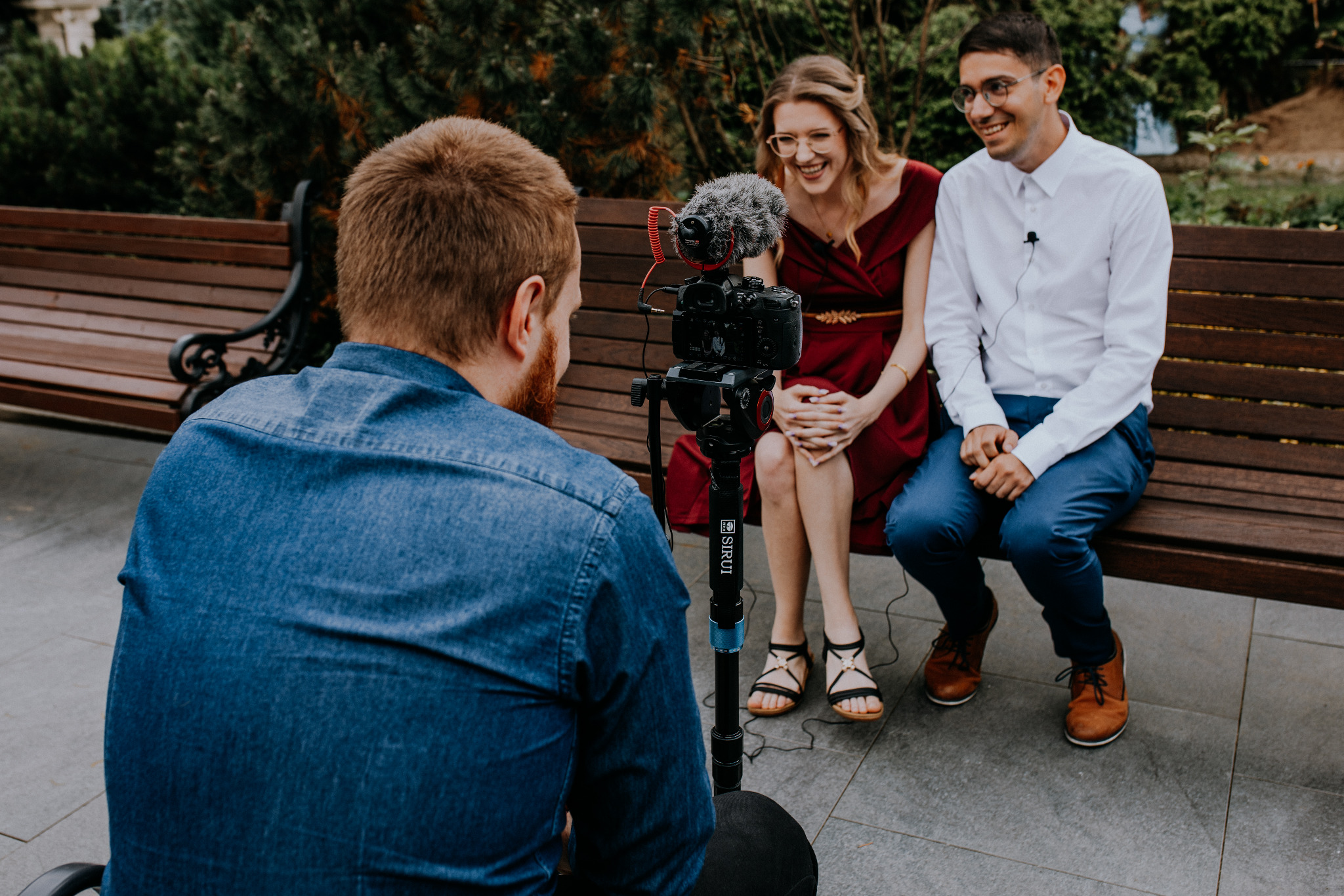 Ujęcie zza ramienia osoby filmującej parę siedzącą na ławce; mikrofon na kamerze, mikroport na koszuli mężczyzny.