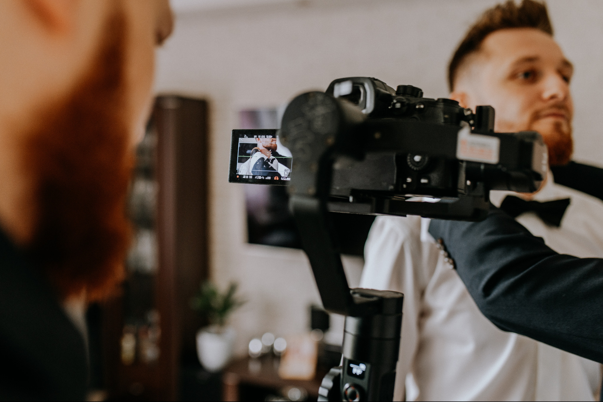 Kamera na stabilizatorze, na ekranie podgląd nagrywanego mężczyzny w garniturze i muszce, w tle drugi mężczyzna z rudą brodą.