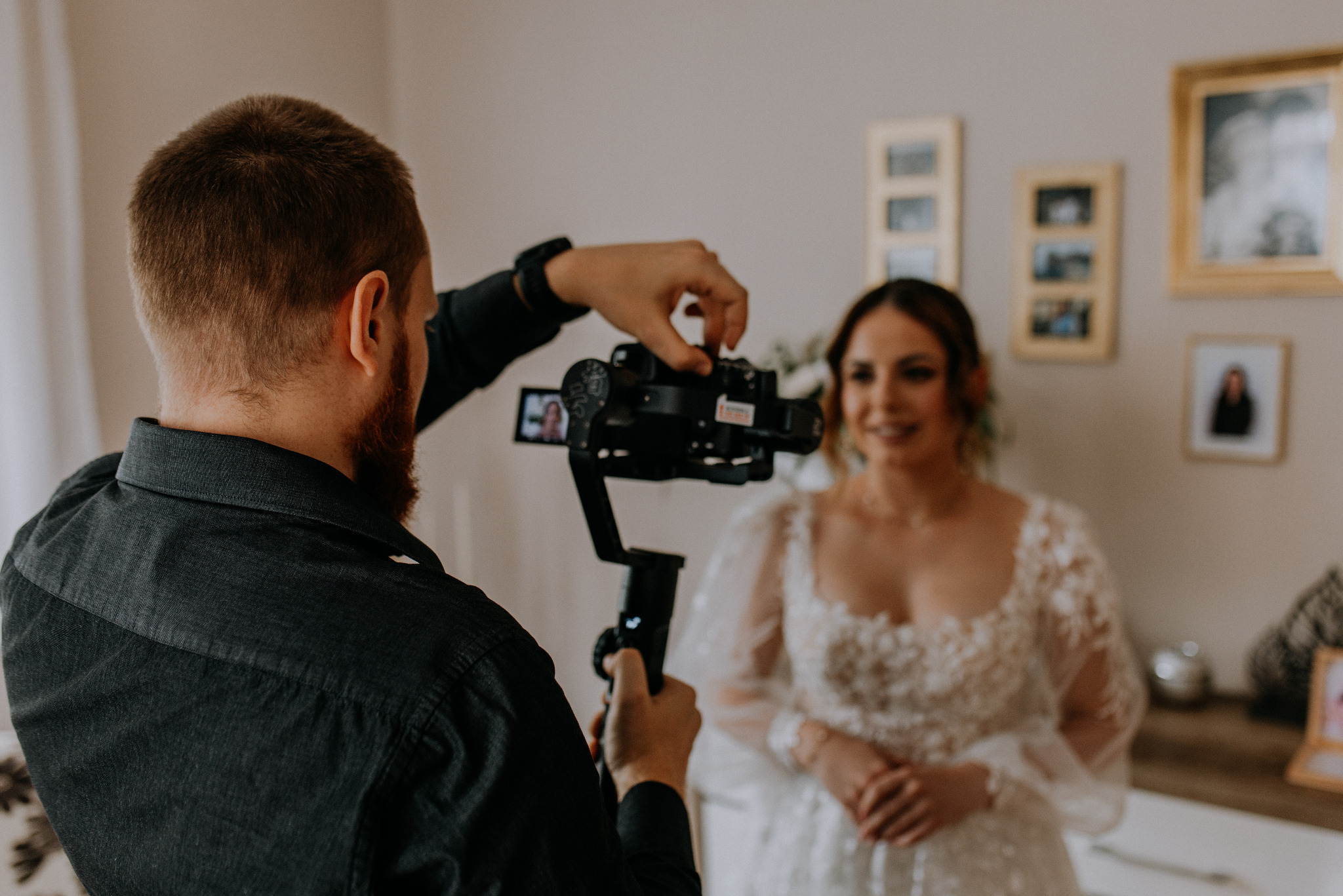 Filmowanie panny młodej w białej sukni ślubnej za pomocą kamery na stabilizatorze, widok zza pleców operatora, zdjęcia w złotych ramkach na ścianie w tle.