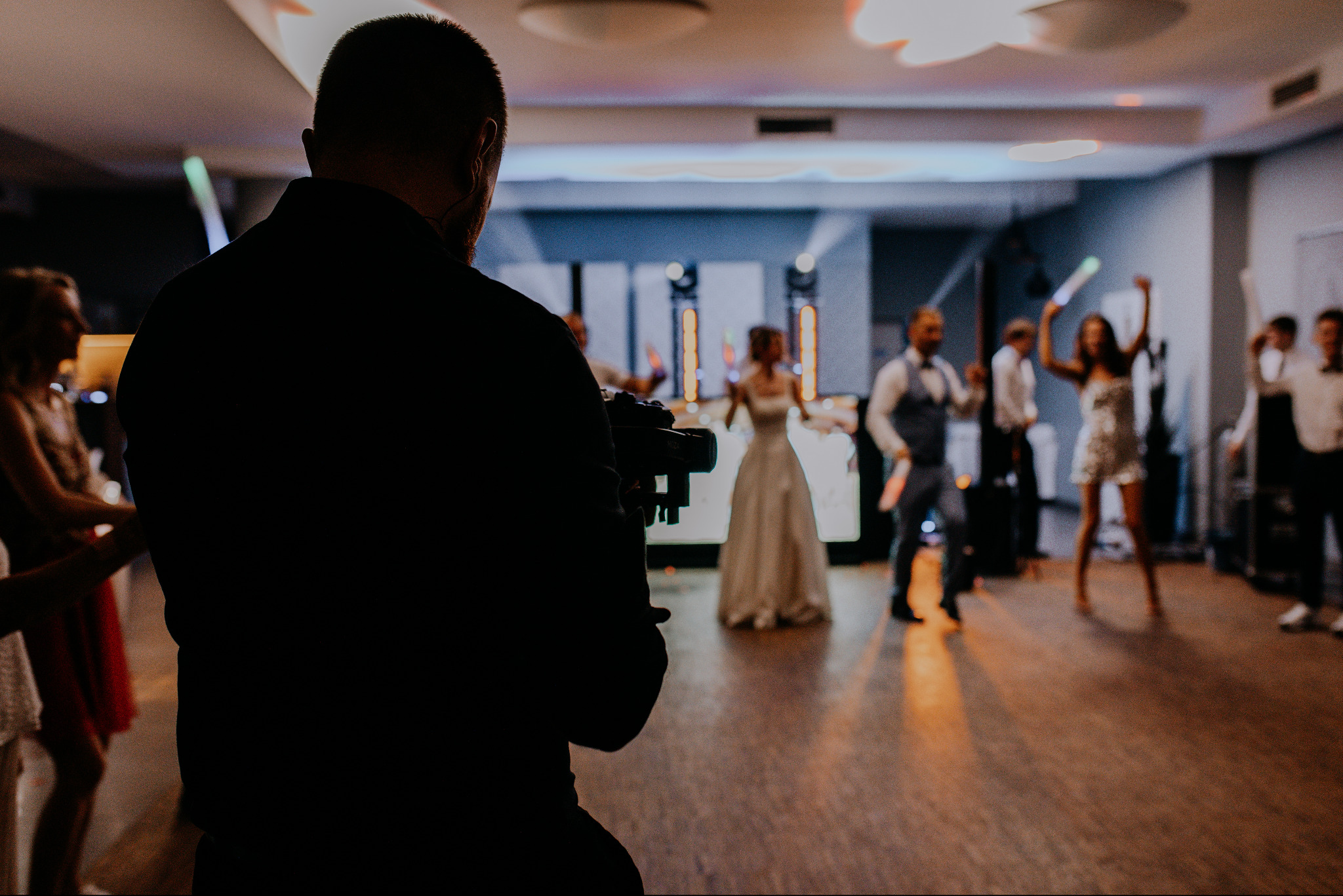 Kamerzysta filmuje parę młodą i gości podczas weselnego przyjęcia, widok zza jego pleców.