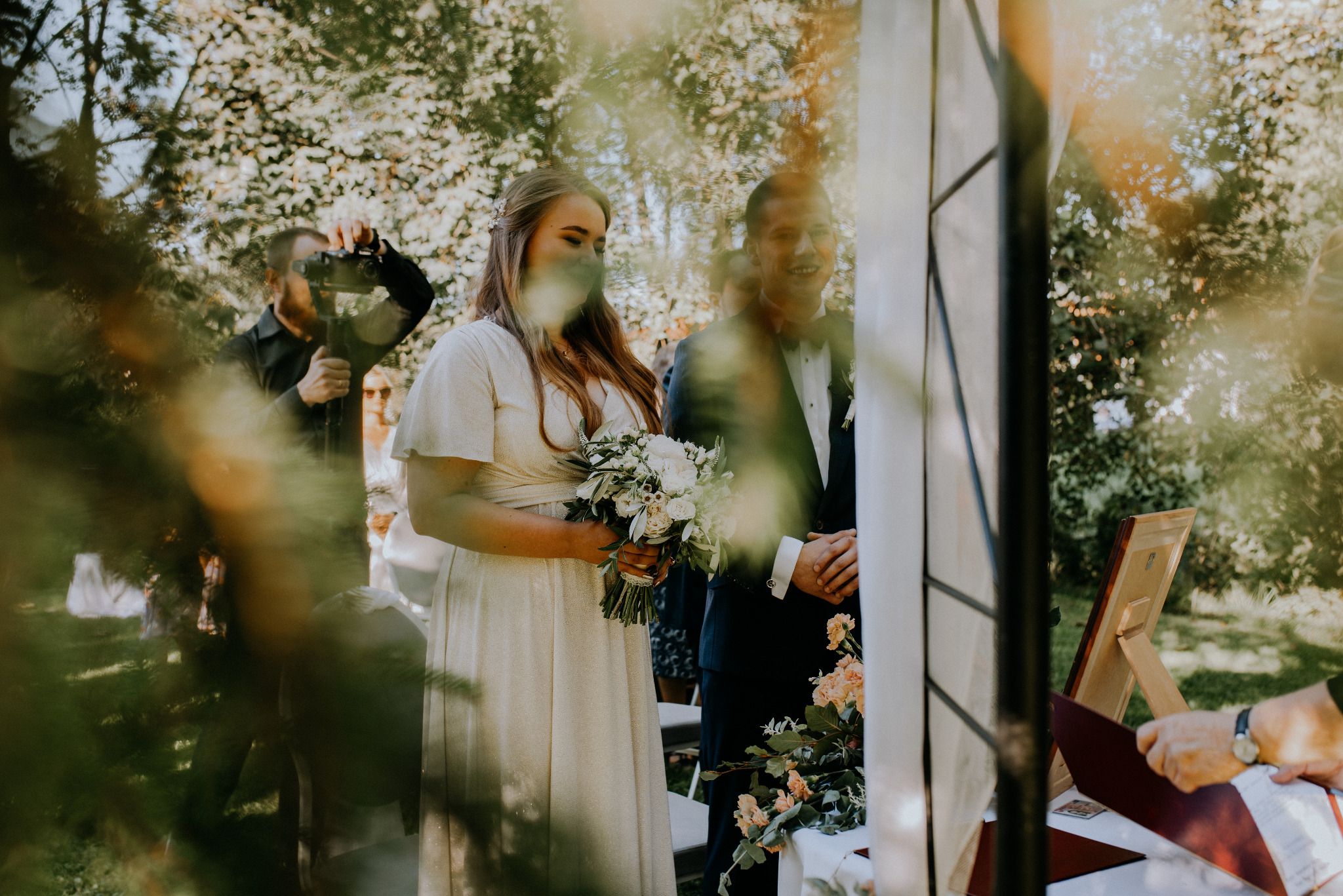 Para młoda podczas ceremonii ślubnej na świeżym powietrzu, w tle kamerzysta filmujący wydarzenie, widoczne elementy dekoracji ślubnej.
