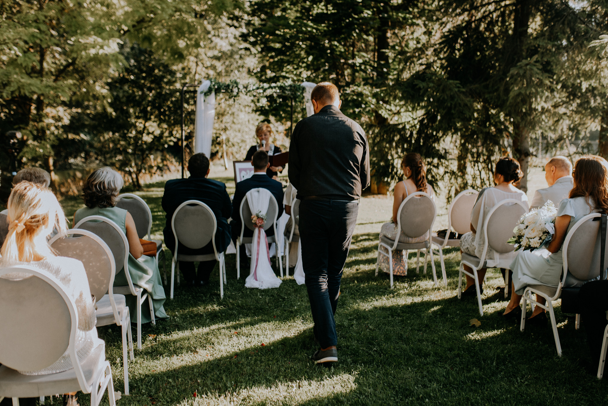 Uroczystość ślubna w plenerze: goście siedzą na krzesłach ustawionych na trawie, w tle widać parę młodą i osobę udzielającą ślubu, dekoracje kwiatowe.