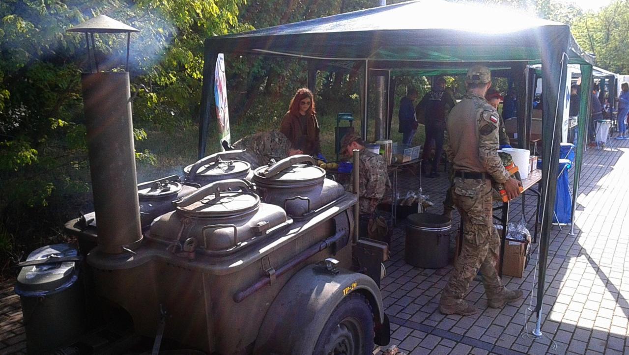 Polowa kuchnia wojskowa z dymiącym kominem, przygotowująca posiłki pod namiotami podczas imprezy plenerowej.