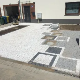 Układanie jasnoszarej kostki granitowej z ciemnoszarymi i białymi obramowaniami, widoczna maszyna do ubijania i pracownicy w tle, częściowo ukończony wzór.