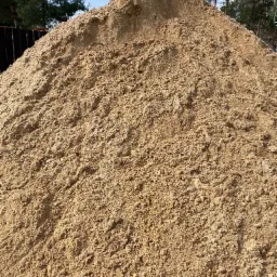 Duża pryzma piasku budowlanego o drobnej granulacji, widoczna na tle ogrodzenia i drzew iglastych pod błękitnym niebem.