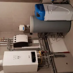 Nowoczesna kotłownia z białym kotłem kondensacyjnym Buderus, szarym zasobnikiem ciepłej wody, niebiesko-białym zmiękczaczem wody oraz rozbudowaną instalacją hydrauliczną z rur miedzianych...