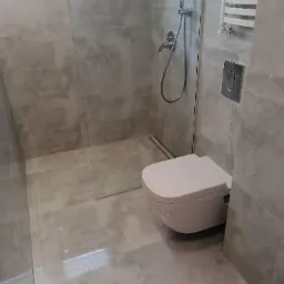 Wykończona łazienka w szarych płytkach imitujących beton, z kabiną prysznicową typu walk-in, podwieszaną toaletą i grzejnikiem drabinkowym.