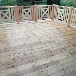 Drewniany taras z jasnego drewna z balustradą o wzorze krzyży, otoczony zielenią drzew.