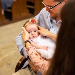 Fotoola - Rodzice trzymają niemowlę w białej sukience i z opaską z kwiatami na głowie, przygotowując się do chrztu w kościele.