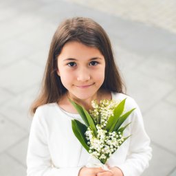 Fotoola - Dziewczynka w białej sukience trzyma bukiet konwalii, uśmiechając się do kamery na tle szarej kostki brukowej.