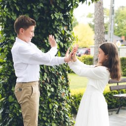 Fotoola - Uśmiechnięte rodzeństwo, chłopiec w białej koszuli i beżowych spodniach oraz dziewczynka w białej sukience, bawią się na tle zieleni w plenerze w Łodzi.