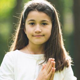 Fotoola - Portret dziewczynki z rękami złożonymi do modlitwy na tle zieleni, z delikatnym uśmiechem, w białej bluzce i z ozdobną opaską na włosach, oświetlona promieniami słońca.