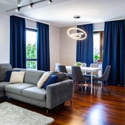 Fotoola - Nowoczesny salon z szarym narożnikiem, białym dywanem, ciemnymi zasłonami i drewnianą podłogą. W tle biały stół jadalny z szarymi krzesłami i designerska lampa.