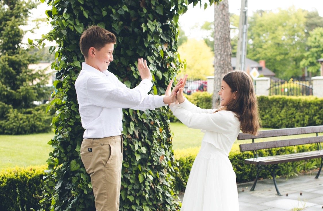 Uśmiechnięte rodzeństwo, chłopiec w białej koszuli i beżowych spodniach oraz dziewczynka w białej sukience, bawią się na tle zieleni w plenerze w Łodzi.