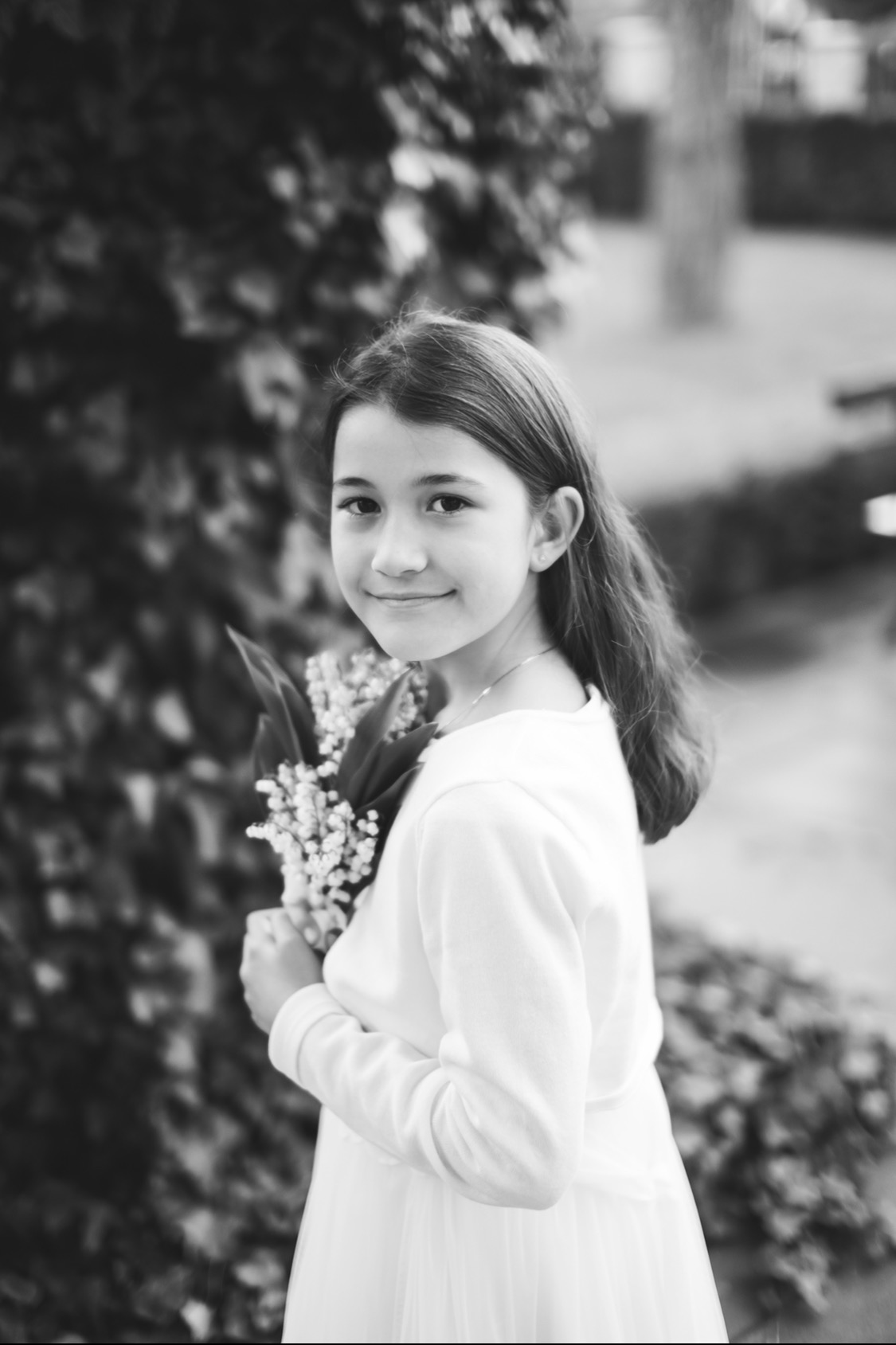 Czarno-białe portretowe zdjęcie uśmiechniętej dziewczynki w białej sukience trzymającej bukiet konwalii na tle zieleni.