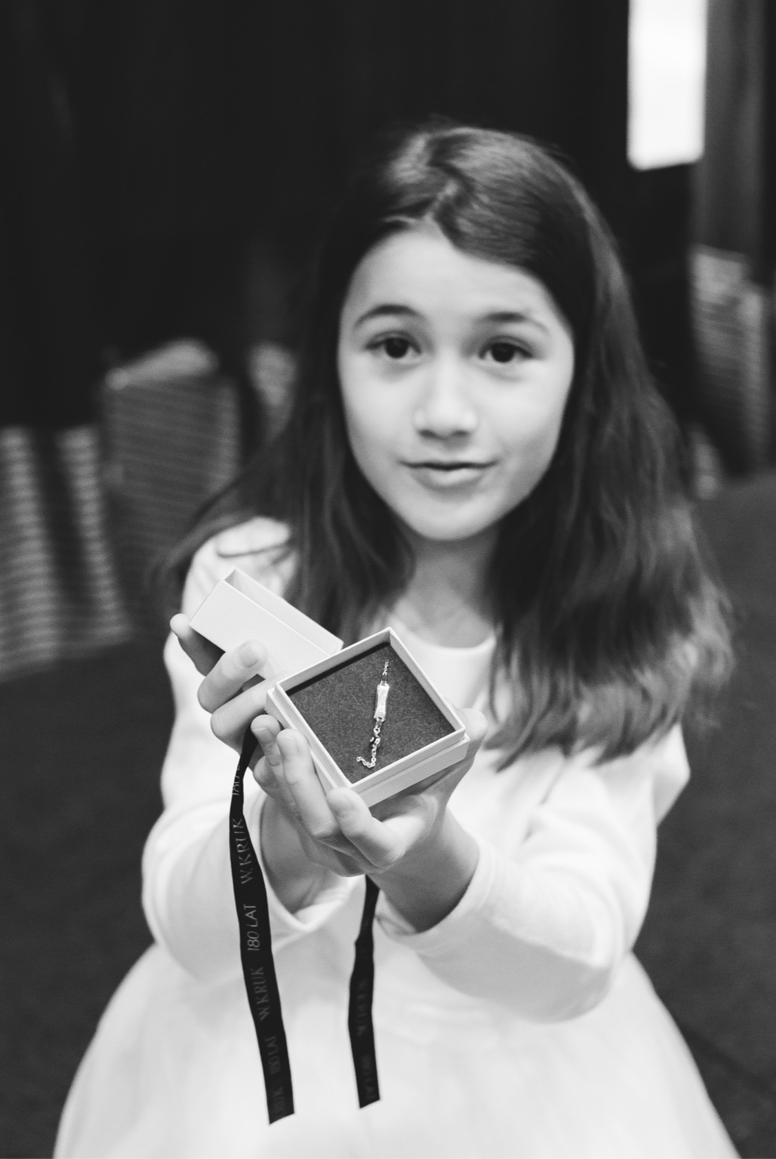 Portret dziewczynki w białej sukience, trzymającej otwarte pudełko z srebrnym wisiorkiem na czarnym tle, pudełko przewiązane wstążką z napisem 'W.KRUK 180 LAT'.