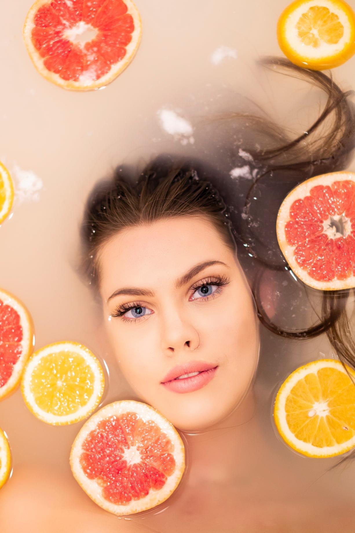 Portret kobiety z jasnymi oczami w mlecznej kąpieli otoczonej plasterkami grejpfrutów i pomarańczy, włosy częściowo zanurzone w wodzie.
