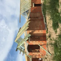 Budowa domu jednorodzinnego w trakcie wznoszenia ścian i konstrukcji dachu z drewnianych belek na tle błękitnego nieba.