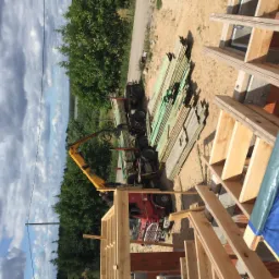 Ciężarówka z żółtym dźwigiem dostarcza drewniane belki na plac budowy domu w trakcie wznoszenia szkieletu, widok z góry na konstrukcję i rozładunek materiałów.