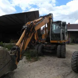 Żółta koparka kołowa marki Liebherr 900 stojąca na piaszczystym placu budowy z widocznym łyżką i kabiną, zadaszenie w tle i pochmurne niebo.