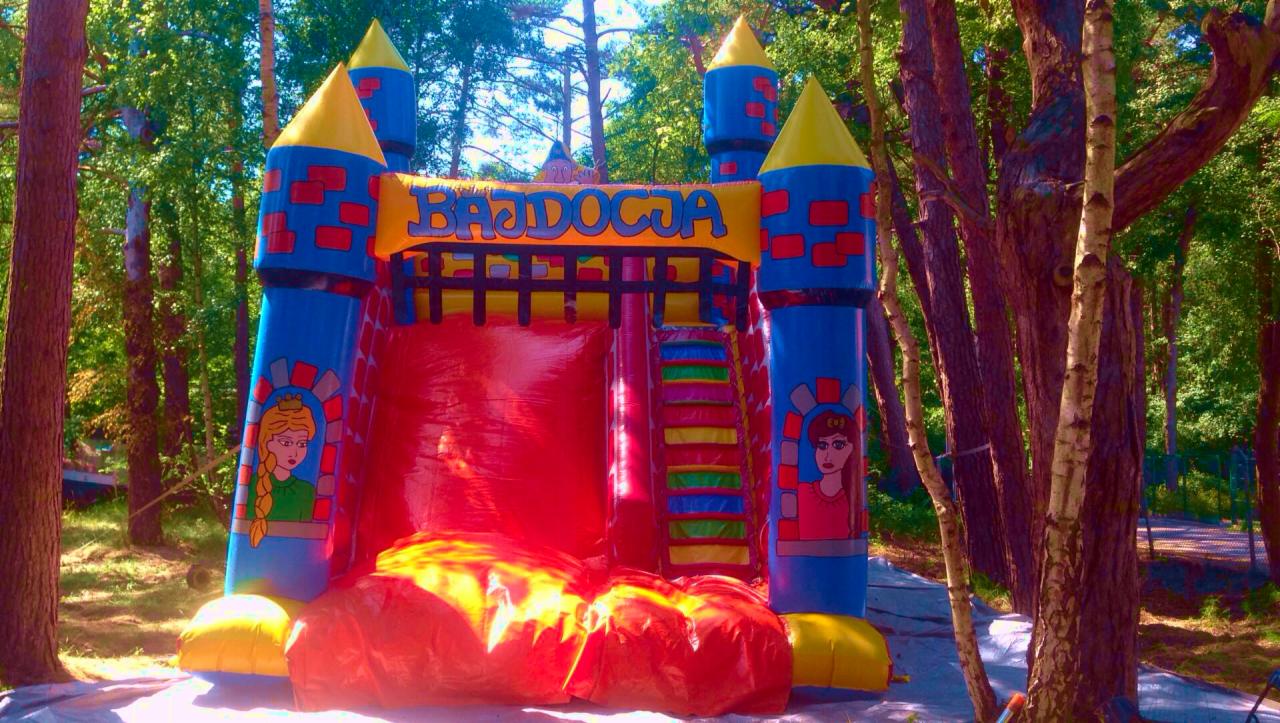 Kolorowy dmuchany zamek 'Bajdocja' z motywami księżniczek, ustawiony w lesie na rozłożonej folii ochronnej.