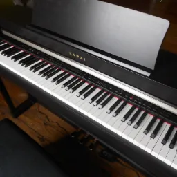 Czarny fortepian cyfrowy Kawai z widocznymi klawiszami i pulpitem na nuty, ustawiony na drewnianej podłodze, z widocznym czarnym stołkiem.