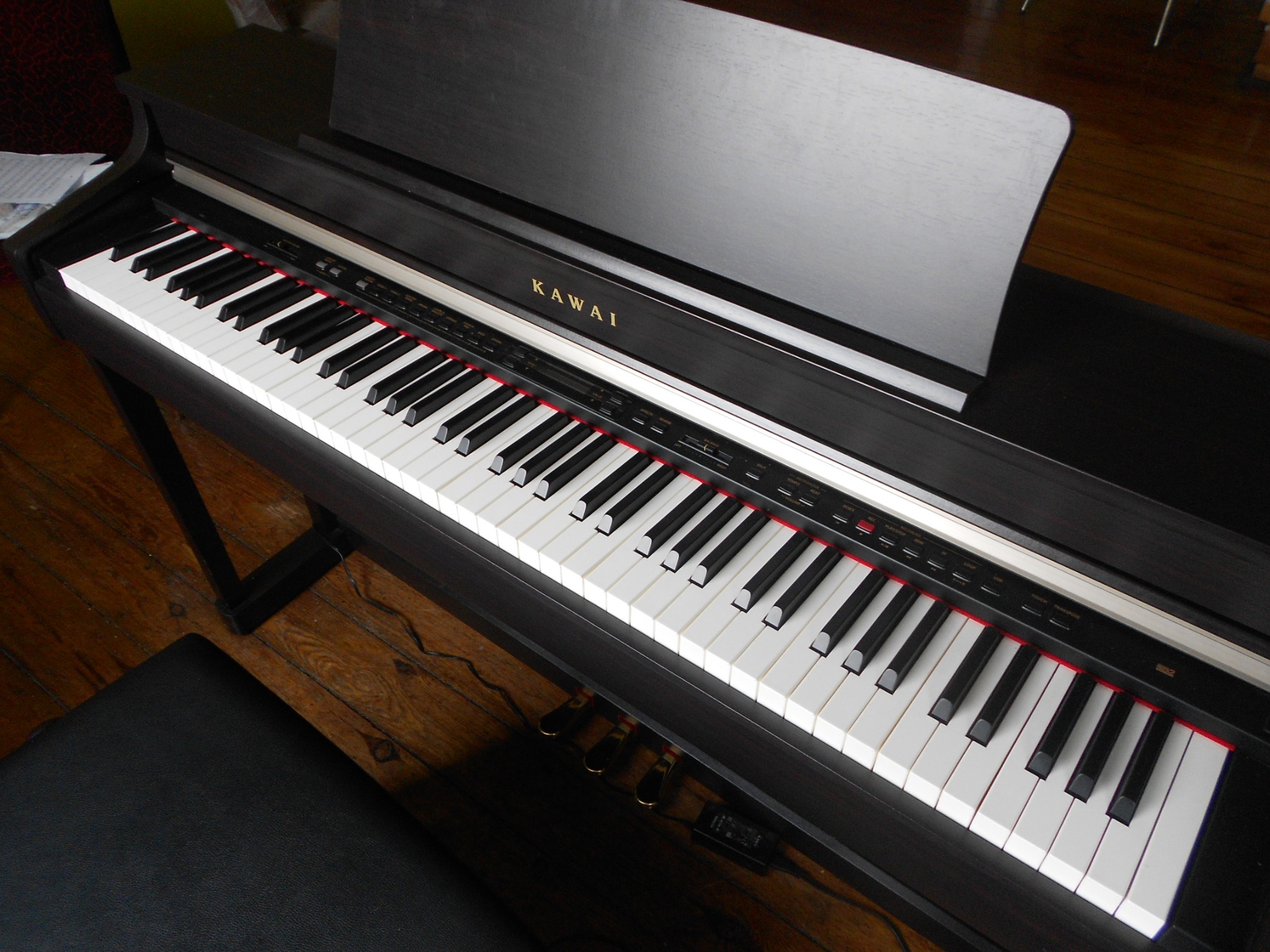 Czarny fortepian cyfrowy Kawai z widocznymi klawiszami i pulpitem na nuty, ustawiony na drewnianej podłodze, z widocznym czarnym stołkiem.