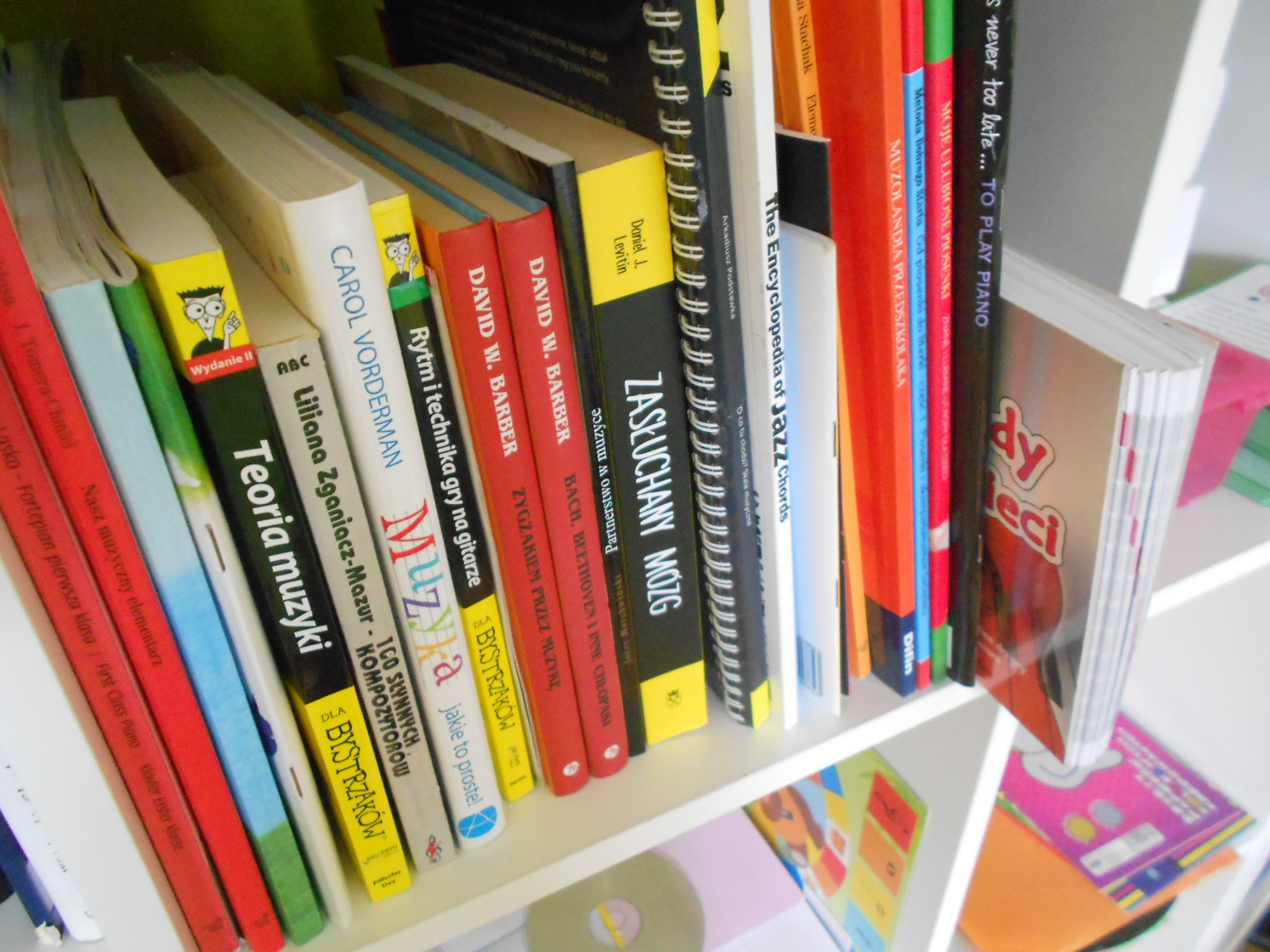 Półka z książkami o tematyce muzycznej, widoczne tytuły dotyczące teorii muzyki, rytmu, gry na gitarze, jazzu i kompozytorów, ułożone obok siebie na białej półce.