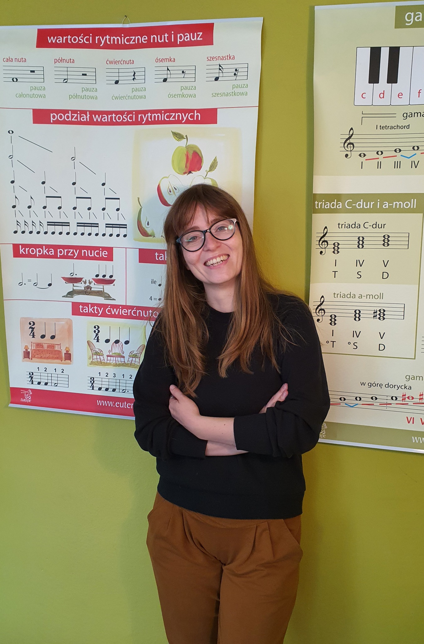 Uśmiechnięta kobieta w okularach stoi na tle ściany z plakatami edukacyjnymi dotyczącymi wartości rytmicznych nut i akordów muzycznych.