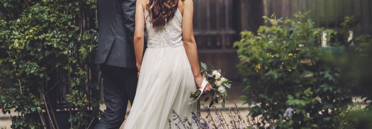 Para młoda, widok z tyłu, idąca obok zielonych krzewów; panna młoda w sukni z cekinową górą i bukietem białych kwiatów.