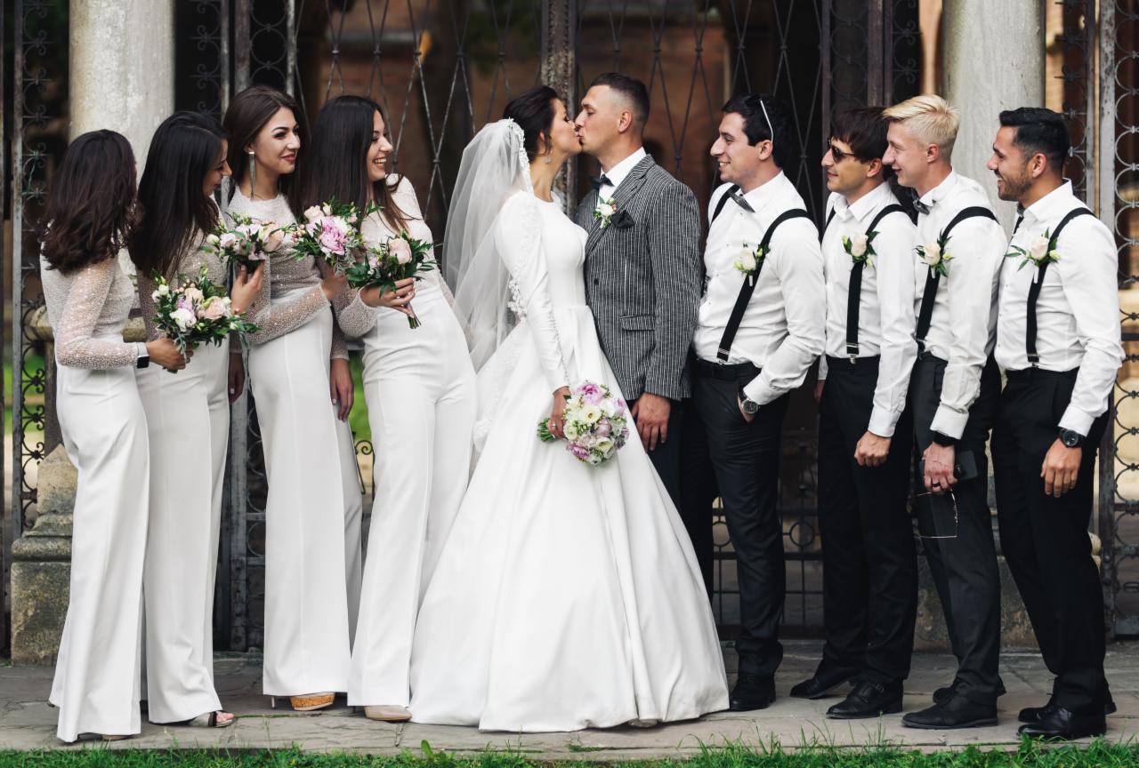 Panna młoda w długiej białej sukni i welonie całuje pana młodego w szarym garniturze na tle bramy z druhnami w białych kombinezonach i drużbami w czarnych spodniach i białych koszulach z szelkami.