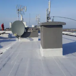 Biała, płaska powierzchnia dachu z widocznymi kominami, antenami satelitarnymi i telekomunikacyjnymi, zabezpieczona hydroizolacją. W tle widoczny pracownik w czerwonym kombinezonie i zabudowa miejska.