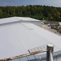 Widok z góry na duży, płaski, jasnoszary dach z blachy trapezowej, z widocznymi świetlikami, w Inowrocławiu. W tle drzewa i zaparkowane pojazdy.