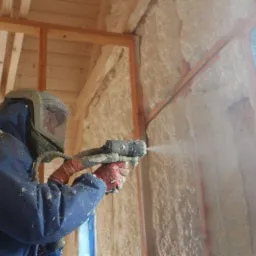 Pracownik w kombinezonie ochronnym aplikuje pianę izolacyjną na drewnianą konstrukcję ściany wewnątrz budynku, widoczne świeżo nałożona warstwa izolacji.