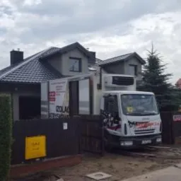 Samochód dostawczy z reklamą materiałów izolacyjnych zaparkowany przed domem z nowym, ciemnym dachem z dachówki ceramicznej i drewnianym ogrodzeniem.