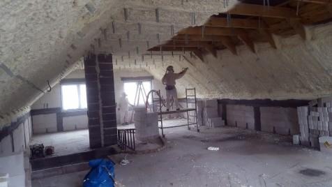 Pracownik w kombinezonie ochronnym aplikuje izolację natryskową na poddaszu, widoczne elementy konstrukcji dachu i ściany z pustaków, część powierzchni już pokryta pianą.