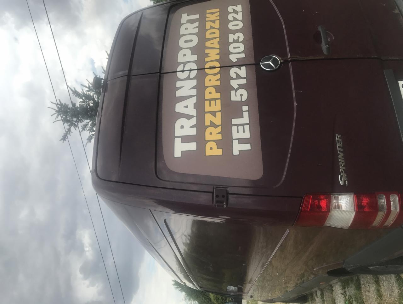 Tył bordowego busa Mercedes Sprinter z naklejonym napisem 'TRANSPORT PRZEPROWADZKI TEL. 512 103 022' na tylnych drzwiach, widziany z boku.