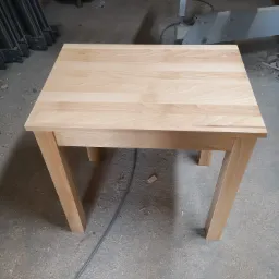 Jasny, drewniany stół o kwadratowym blacie i prostych nogach, widoczny w warsztacie stolarskim, z maszyną do obróbki drewna w tle.