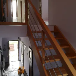 Drewniane schody z metalowymi barierkami w trakcie montażu, widoczny bałagan i narzędzia w korytarzu, surowy stan wykończenia ścian na górze.