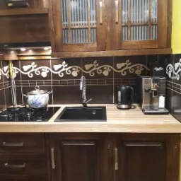 Ciemnobrązowa zabudowa kuchenna ze szklanymi witrynami, czarnym zlewem i jasnym blatem w Śmiglu, uzupełniona o czajnik elektryczny i ekspres do kawy.