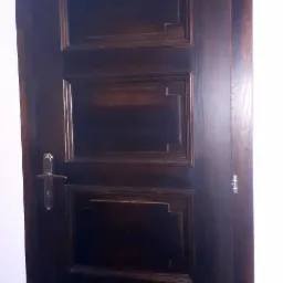 Ciemnobrązowe, drewniane drzwi z trzema panelami o geometrycznych zdobieniach, widoczne zawiasy i klamka.