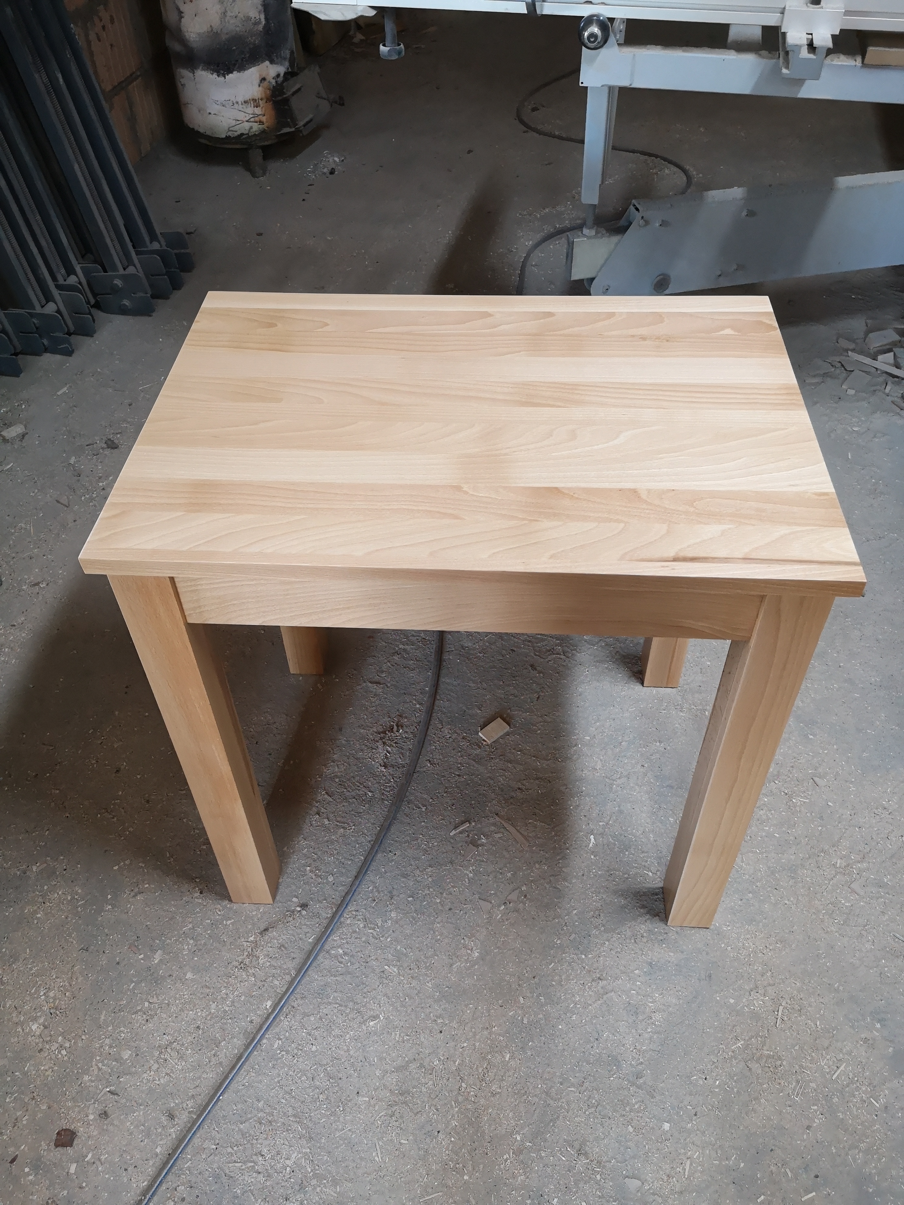 Jasny, drewniany stół o kwadratowym blacie i prostych nogach, widoczny w warsztacie stolarskim, z maszyną do obróbki drewna w tle.