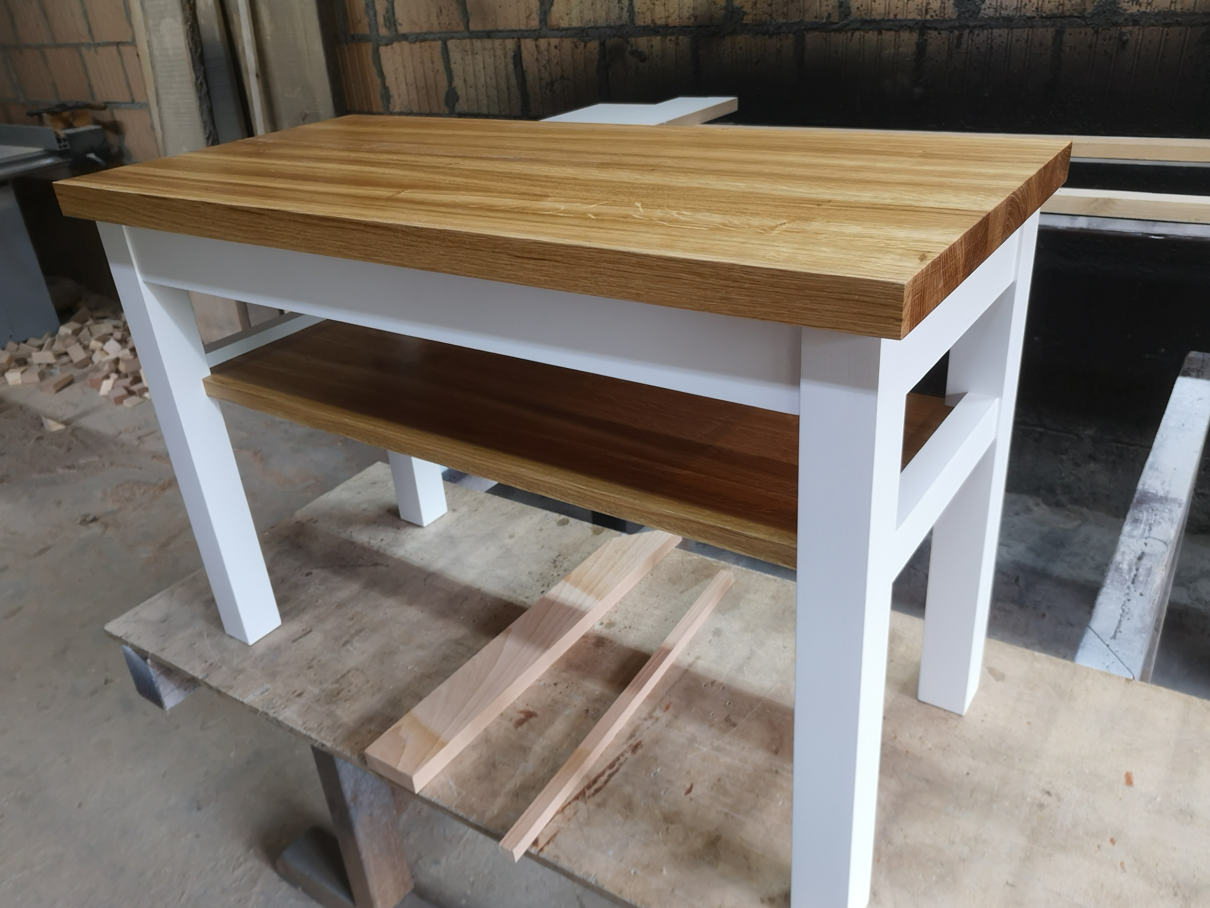 Stolik kawowy z blatem z jasnego drewna i białymi nogami, dodatkowa półka z ciemniejszego drewna, widoczne dwie listwy na stole warsztatowym w tle ceglana ściana.