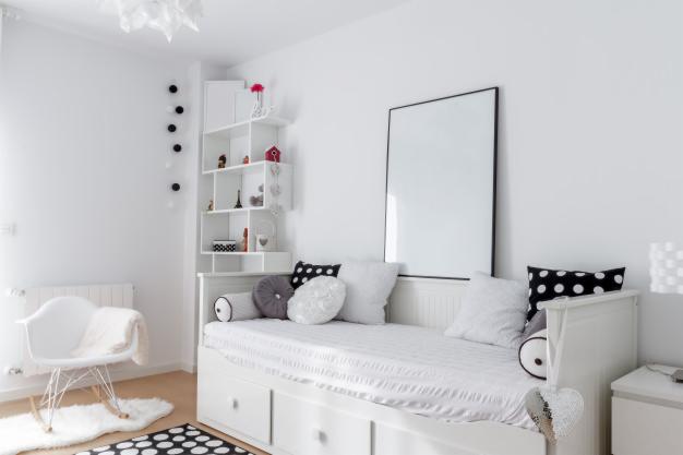 Biały pokój dziecięcy z łóżkiem typu daybed, regałem narożnym i krzesłem bujanym na tle jasnej podłogi.