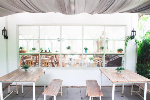 Jasne wnętrze patio z drewnianymi stołami i ławkami, widok na ogród przez duże okno z białymi zasłonami, oświetlone lampami ściennymi w stylu vintage.