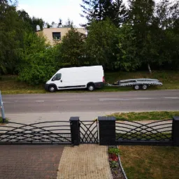 Biały bus z lawetą na poboczu drogi, widok zza ozdobnego ogrodzenia. Laweta pusta, gotowa do załadunku. Tło: zieleń drzew i budynek z oknem.