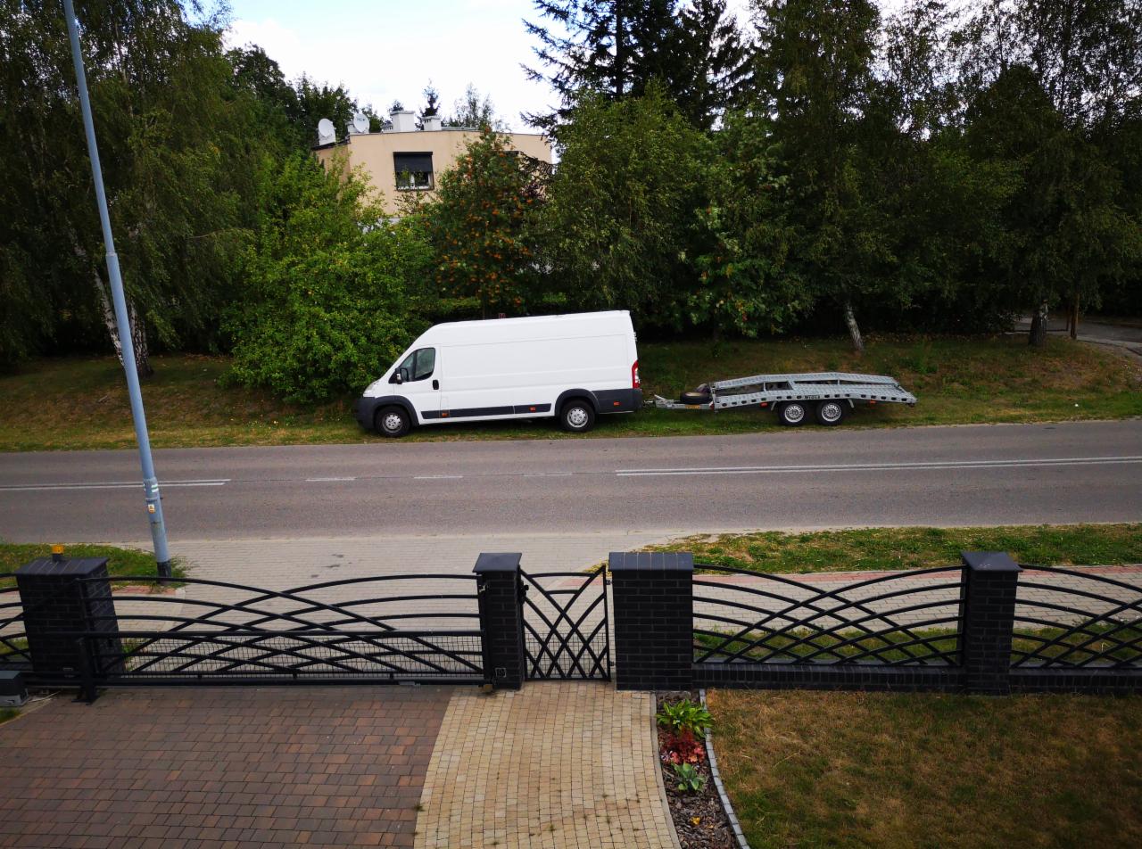 Biały bus z lawetą na poboczu drogi, widok zza ozdobnego ogrodzenia. Laweta pusta, gotowa do załadunku. Tło: zieleń drzew i budynek z oknem.