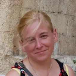 Portret uśmiechniętej kobiety z blond włosami na tle kamiennej ściany, z delikatnym łańcuszkiem na szyi i paskiem torby na ramieniu.