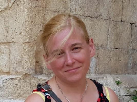 Portret uśmiechniętej kobiety z blond włosami na tle kamiennej ściany, z delikatnym łańcuszkiem na szyi i paskiem torby na ramieniu.